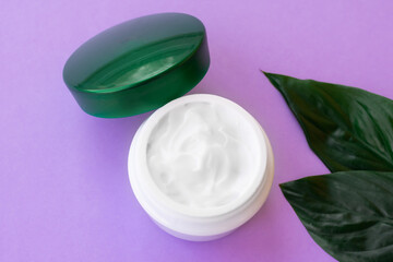 Obraz na płótnie Canvas Soft white cream in jar with bright green leaves on purple background. Spa treatment and procedure at home.