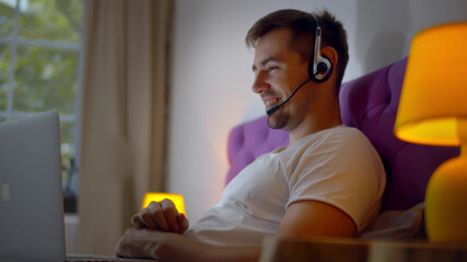 Handsome young man using laptop and headset for video call lying in bed