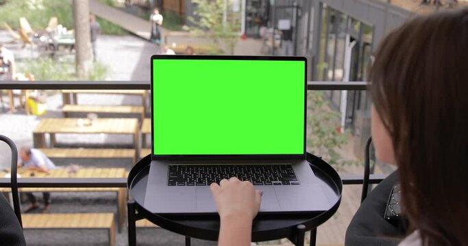 Young Woman at Coffee Shop Working on a Laptop MacBook Pro Computer with Green Mock-up Screen. She's Sitting On a Bean Bag Chair in Coworking open space outdoor office. Over the Shoulder Camera Shot.