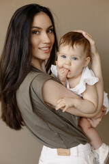 Young smiling woman mom is holding a small cute baby girl in the studio on beige background isolated. Happy motherhood, lifestyle