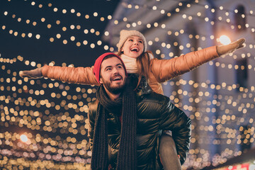 Photo of positive couple having fun christmas x-mas around evening outside illumination boyfriend piggyback girlfriend holding hands fly