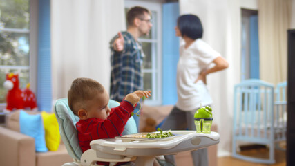 Cute little boy sitting in highchair with parents arguing on background