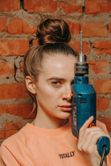 A beautiful girl with a drill stands against a brick wall background. Holds the drill like a gun