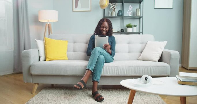 Happy African American curly female typing on tablet while sitting on couch and smiling in living room. Cheerful beautiful girl with smile on face browsing on device at home. Leisure concept