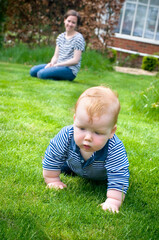 Proud mother watching young baby boy learning to crawl on the grass in the garden. Portrait format.