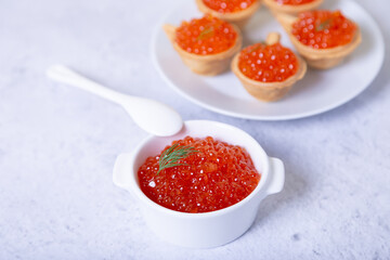 Red caviar (salmon caviar) in a white cup. In the background is a plate with tartlets and caviar. Selective focus, close up.