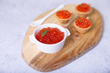 Red caviar (salmon caviar) in a white bowl on a wooden board. In the background are tartlets with caviar. Selective focus, close-up.