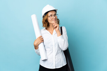 Young architect Georgian woman with helmet and holding blueprints over isolated background thinking an idea while looking up