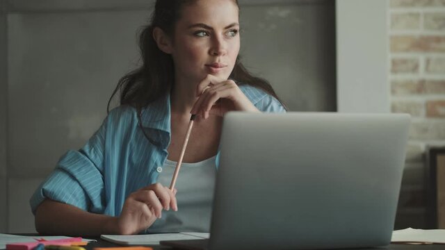 An attractive young woman is working using her laptop computer sitting at the table at home