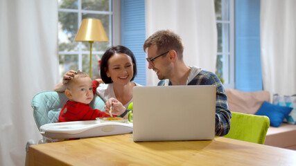 Happy young couple feeding baby boy in highchair and using laptop at home