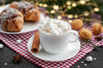 Obraz na płótnie Canvas Christmas breakfast. Christmas morning. A cup of coffee or cocoa with marshmallows and cinnamon and croissants on a dark table with lights. Selective focus. The atmosphere of Christmas. 