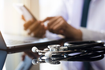 Doctor man hand using mobile smartphone with laptop computer and medical stethoscope on the desk at office. Medical online networking, telehealth, telemedicine, ehr, emr concept