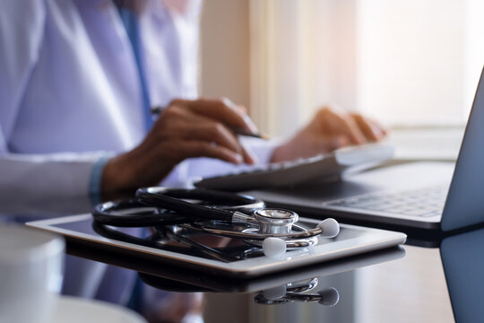 Doctor Or Practitioner Using Calculator And Work On Laptop Computer With Medical Stethoscope And Notebookon The Desk At Clinic Or Hospital. Medical Healthcare Costs ,fees And Revenue Concept.