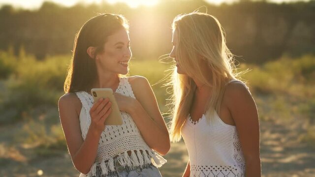 Two laughing girls are looking photos on the smartphone on the beach
