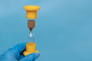 A hand in a blue protective glove holds an hourglass on a blue background, close-up, copy space