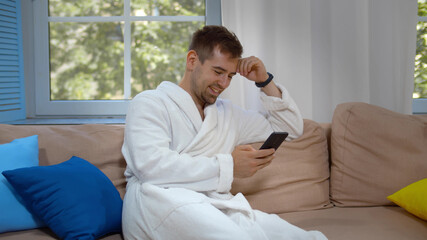 Attractive smiling young man wearing bathrobe sitting on couch at living room using mobile phone
