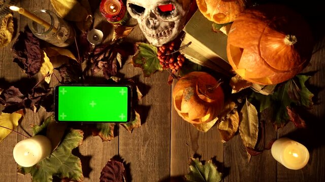 Gestures for using a vertical smartphone with a green screen on background of Halloween. Top view objects for all saints day and phone. Mystical tradition in harvest autumn decor, copy space template