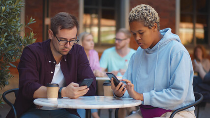 Fototapeta na wymiar Attractive diverse couple in love drinking coffee sitting at cafe table outdoors using mobile phone.