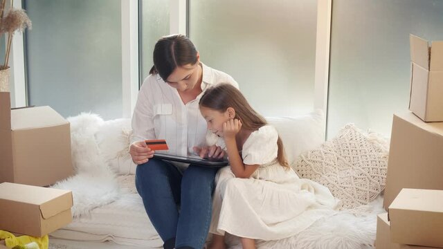 Mother With Her Daughter Spending Money Together at Home. The Mother With a Child Uses a Credit Card for Online Shopping. Purchase Confirmation by the Internet. Uses a Tablet For Online Shopping.