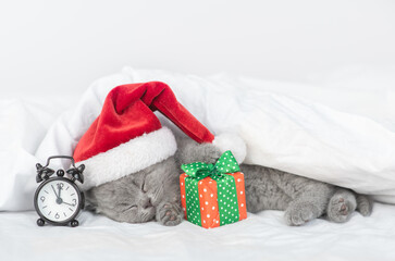 Gray kitten wearing red santa's hat sleeps under blanket with  alarm clock and gift box
