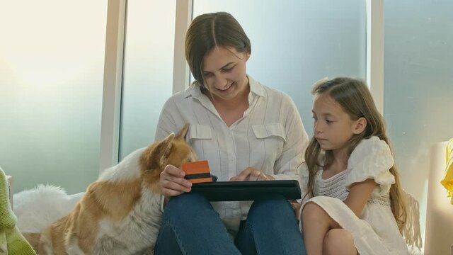 Mother With Her Daughter Spending Money Together.The Mother, a Child and Their Dog Uses a Credit Card for Online Shopping.Purchase Confirmation by the Internet.Uses a Tablet For Online Shopping.