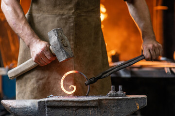 Blacksmith is processing a hot metal object of a spiral shape at anvil in a workshop