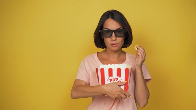 middle aged woman with brown hair stand with some popcorn box on the yellow background. Girl slowly eating popcorn. Woman watch film or movie . High quality 4k footage