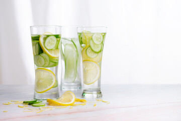 Three glasses of cucumber and lemon refreshing drink decorated with sliced lemon and cucumber on marble table with white background.