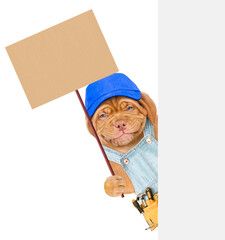 Smiling puppy wearing overalls, blue cap and tool belt holds empty placard in it paw behind empty white banner. isolated on white background