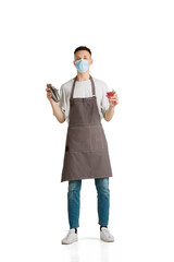 Shaker and drink. Portrait of a young male caucasian barista or bartender in brown apron. Studio white background, copyspace. Working in protective face mask. Professional occupation, drink, service.