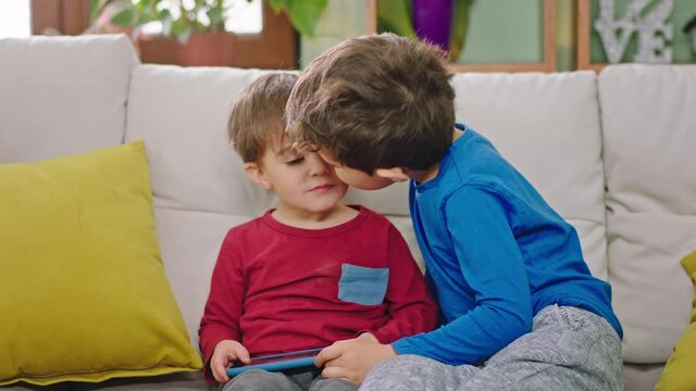Charismatic two kids small boys in front of the camera with a funny face playing a game on their smartphones they giving a kiss then continue to play the game while sitting on the sofa