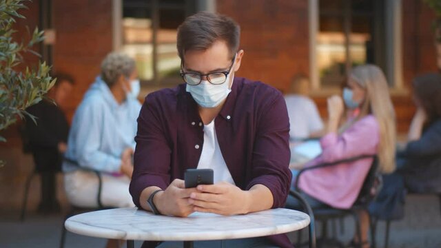 Young guy wearing facial mask surfing internet on mobile phone in outdoors cafe