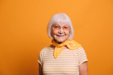 Senior gray-haired woman wearing orange glasses, yellow cravat, striped white and yellow T-shirt standing isolated over orange background and smiling.