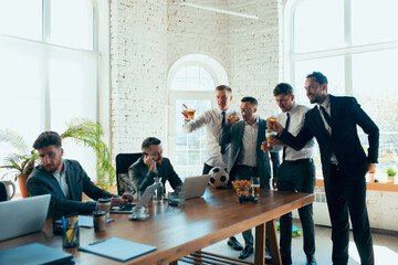 Watching football, drinking beer, cheering. Happy carefree colleagues having fun in office while their co-workers working hard and highly concentrated. Concept of fun, resting, professional occupation