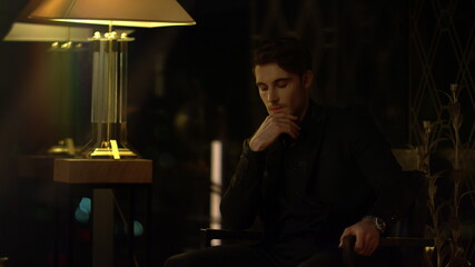 Beautiful man pondering in armchair. Elegant guy touching chin indoors.