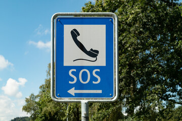emergency phone sign painted with black phone on white-blue background against blue sky and green forest, emergency calls are important to save lives, by day