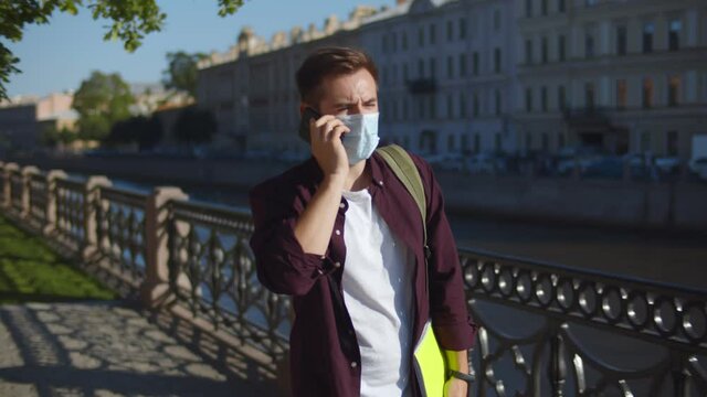 Male student wearing face mask walking outdoors talking on mobile phone