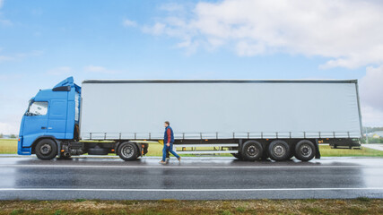 Truck Driver Crosses the Road in the Rural Area and Gets into His Blue Long Haul Semi-Truck with Cargo Trailer Attached. Logistics Company Moving Goods Across Countrie Continent. Side View Shot