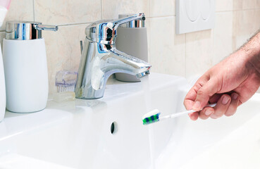 A male hand holding a toothbrush with toothpaste in a white ceramic sink of a bathroom