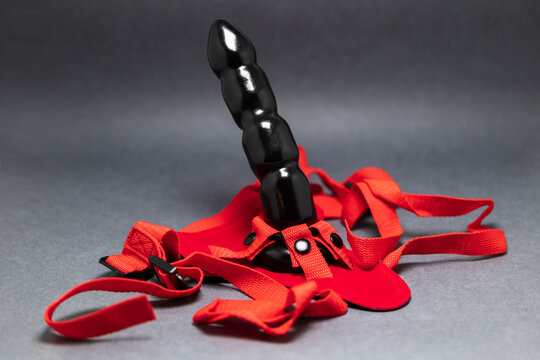 sex toys, black ribbed didlo and red textile strapon on a gray background close up