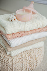 A stack of knitted sweaters, cotton, a candle in a glass jar. Women's sweaters lie on the ottoman. Cozy autumn clothes.