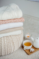 A stack of knitted sweaters, a mug of hot tea, a teapot, a wooden stand on an ottoman. Cozy autumn. Winter breakfast. Roses. Candle.