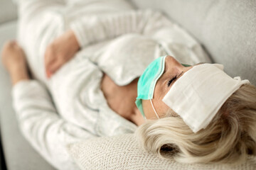 Sick mature woman with face mask having fever and lying down at home.