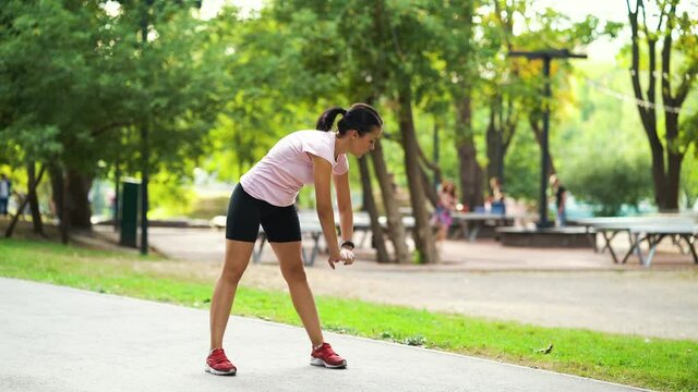 Young woman in sportswear warming up before running in park, stretching legs and arms. Active female training outside. Concept of sport