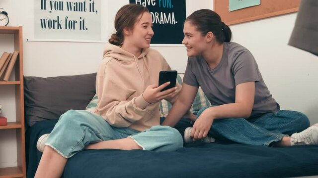 Handheld shot of happy young girls sitting together on bed in their college dorm and chatting while laughing and looking at mobile phone