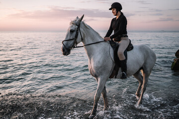 young woman rider in uniform riding a white horse on the beach near the sea