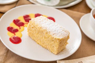 a slice of napoleon cake on a white plate on a served table. In the background there is a kettle, tea, juice.