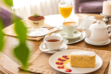 a slice of napoleon cake on a white plate on a served table. In the background there is a kettle, tea, juice.