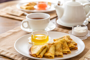 a few pancakes on a plate, honey in a gravy boat. In the background there is a set table, on the table there is a teapot, a cup of tea. Breakfast with pancakes.