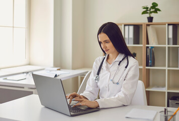 Young female doctor typing on laptop keyword surfing net in modern hospital office